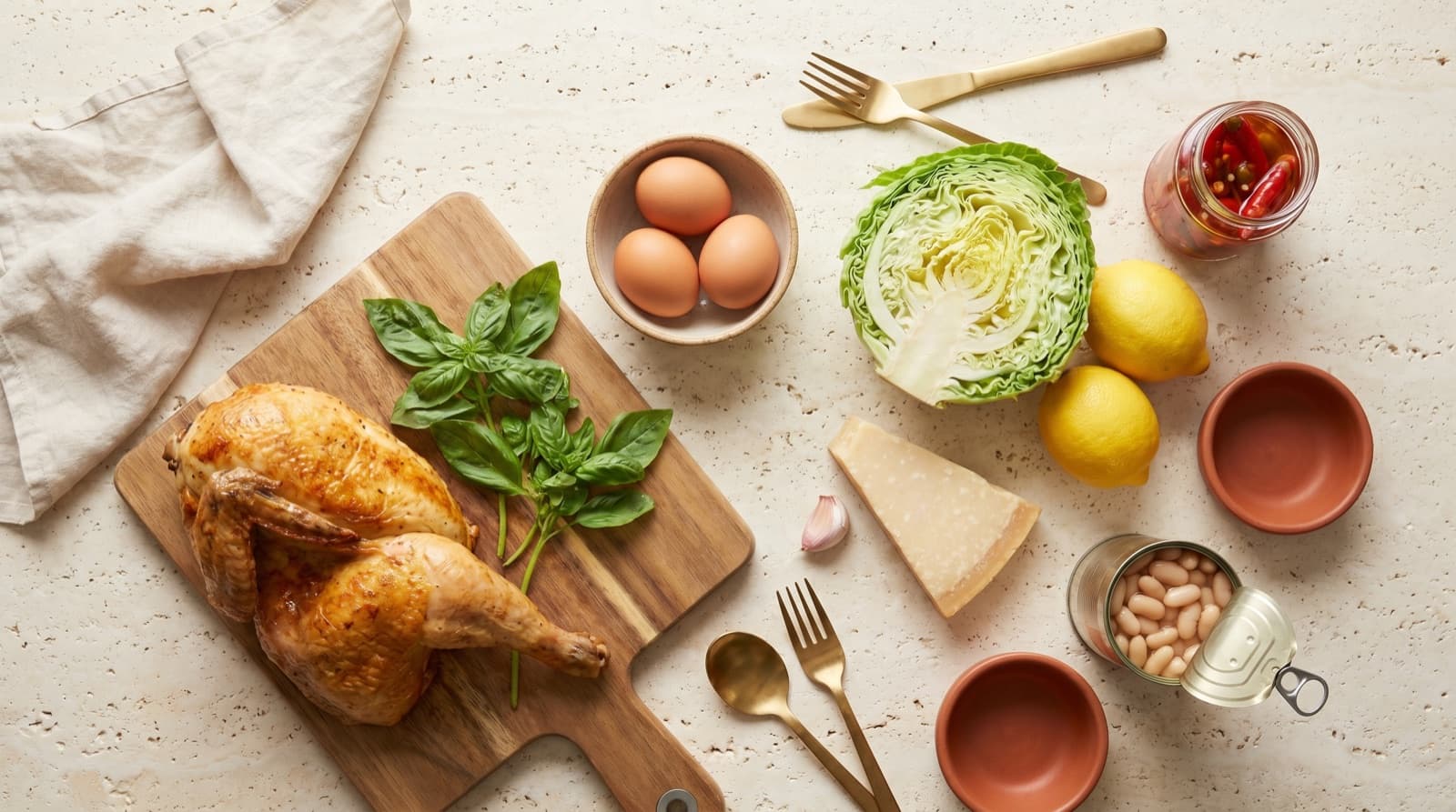 An overhead inventory shot of random fridge-and-pantry ingredients, half a roast chicken on a wooden board, fresh basil, lemons, halved cabbage, eggs, garlic, pickled chillies, beans and parmesan on cream linen