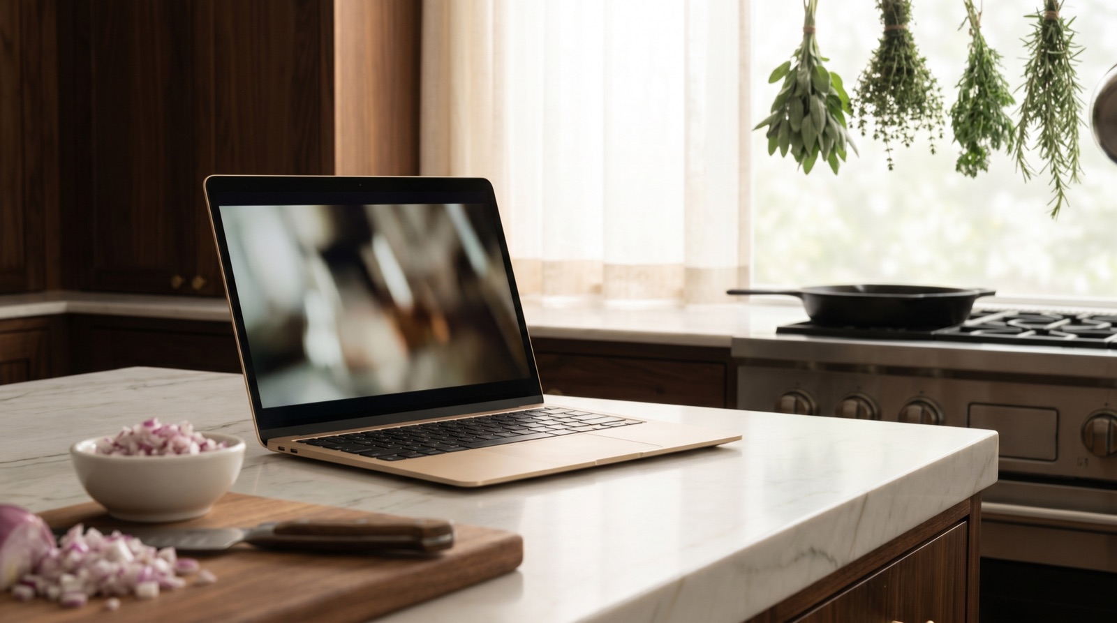 A slim laptop on a marble kitchen counter showing a cooking video, with chopped onions and a chef's knife on a wooden board in the foreground, herbs hanging in a softly blurred background